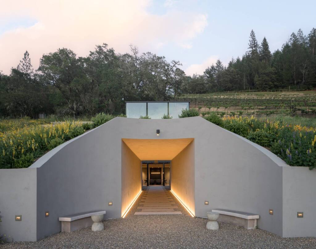 Warmly lit wine cave entrance, with flowers and trees visible above.