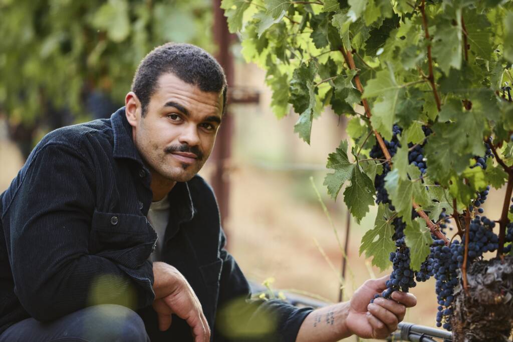 Derek kneeling in the vineyard, holding grapes on the vine