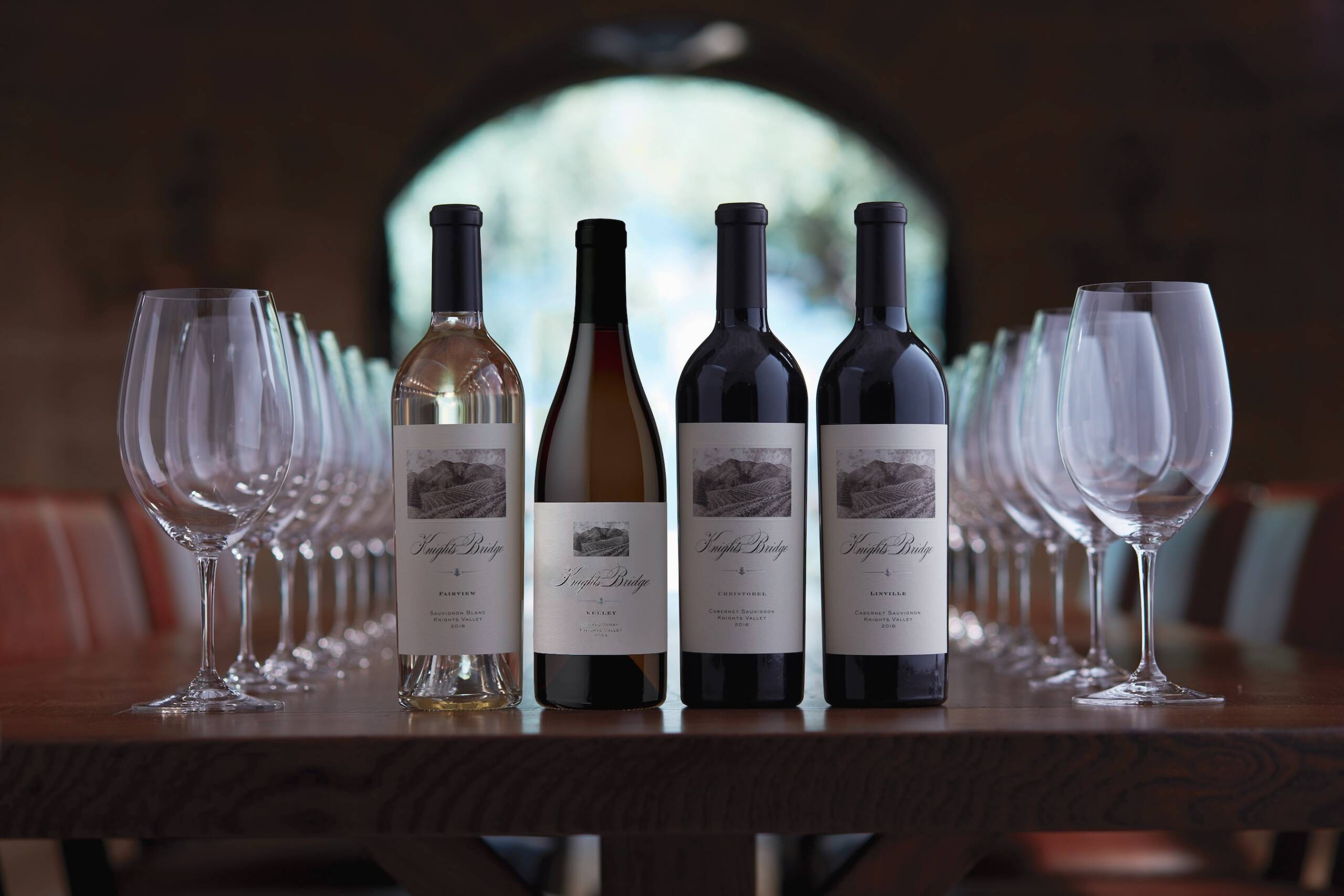 wine bottles and glasses lined up on table with open winery door in background showing outdoors
